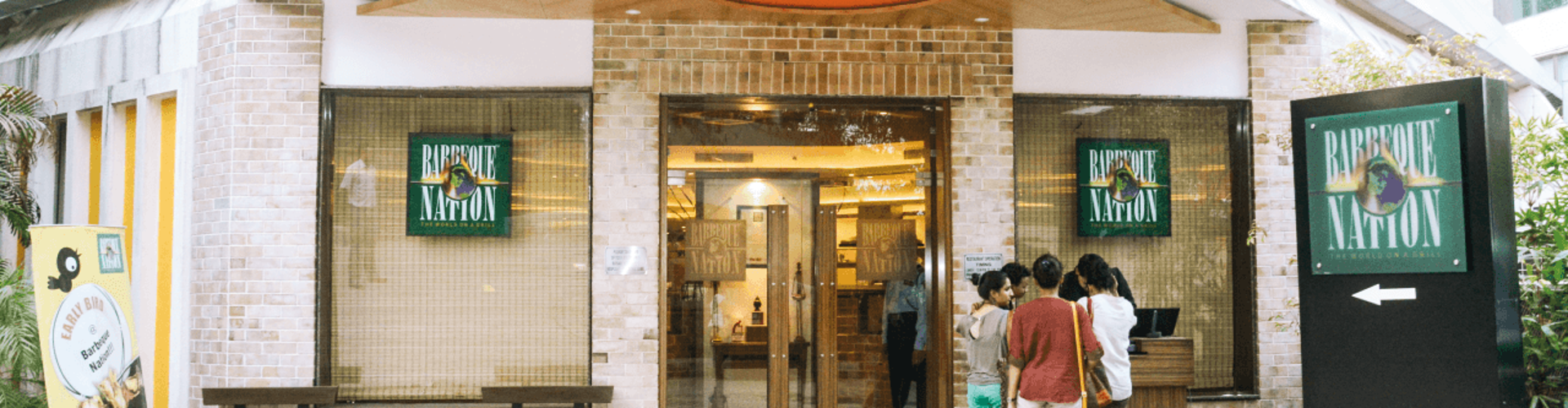 The entrance of a Barbecue Nation restaurant with a few people standing outside, and signage displaying the restaurant‘s name.