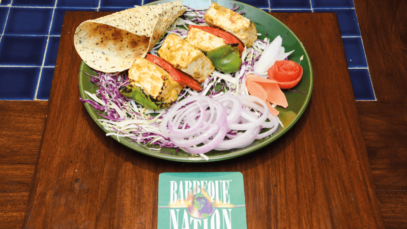 A plate with grilled paneer skewers, onions, salad, and a side of bread, with a Barbeque Nation logo on the placemat.