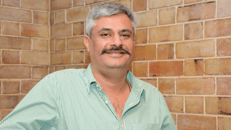A man with gray hair and a mustache, wearing a green striped shirt and standing in front of a brick wall, smiling.