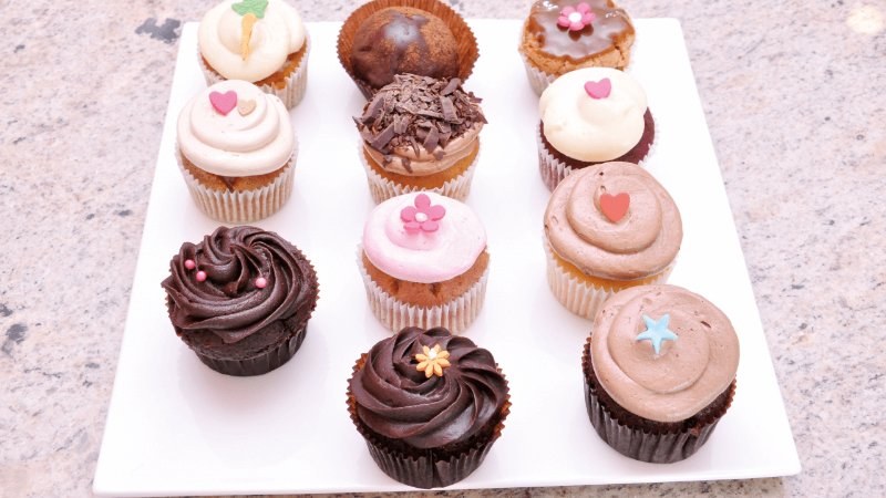 Assorted cupcakes displayed on a white plate, featuring various frosting designs and colorful decorations.