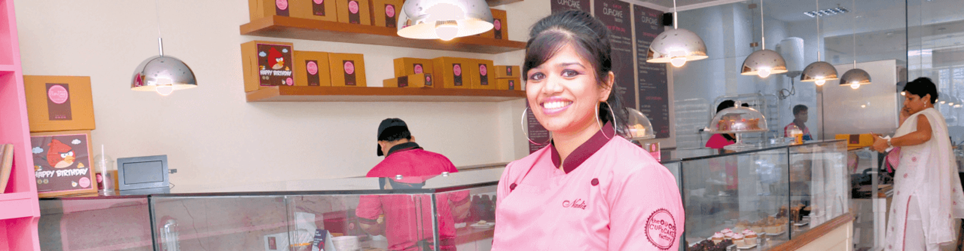 Baker smiling and holding a tray of cupcakes in a colorful bakery with a glass display case.