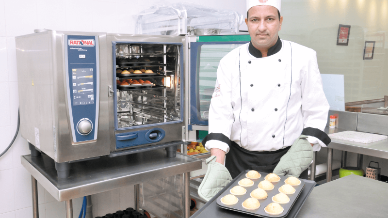 Chef in a kitchen holding a tray of freshly baked cupcakes next to a Rational oven.