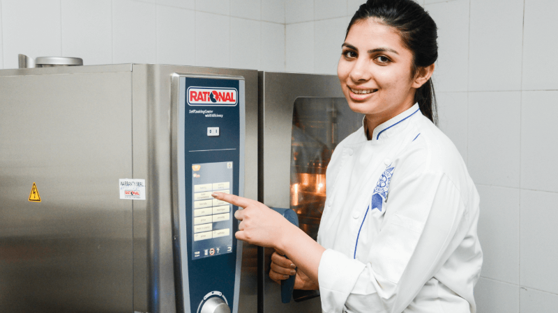 Chef smiling and pointing at the control panel of a Rational oven in a kitchen setting.