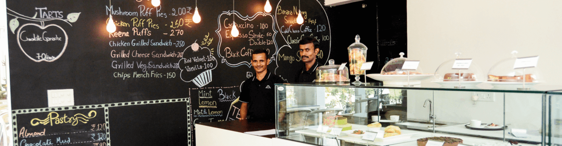 Café interior featuring a chalkboard menu and two staff members standing behind the counter with a display of desserts.