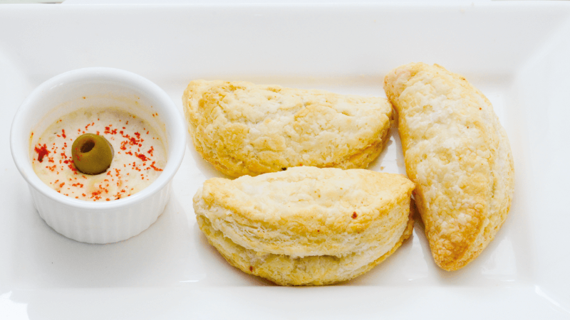 Three golden pastry pockets served on a white plate with a small bowl of dip topped with an olive and sprinkled with red spice.