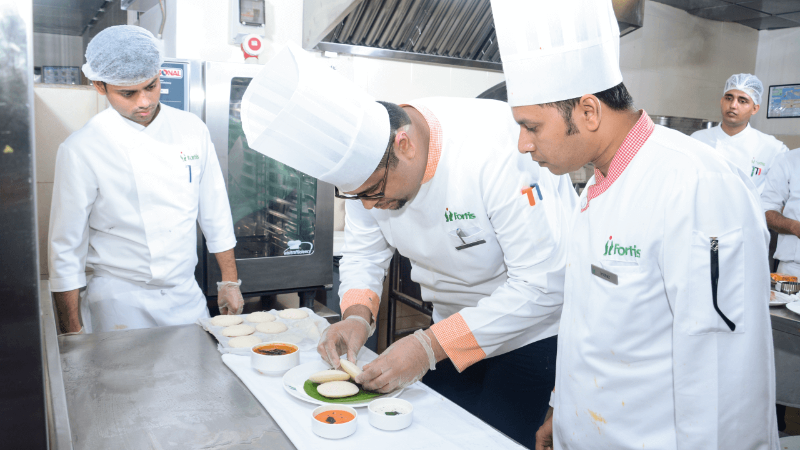 A group of chefs in a kitchen, one chef is plating food while others observe, with dishes and a Rational oven in the background.