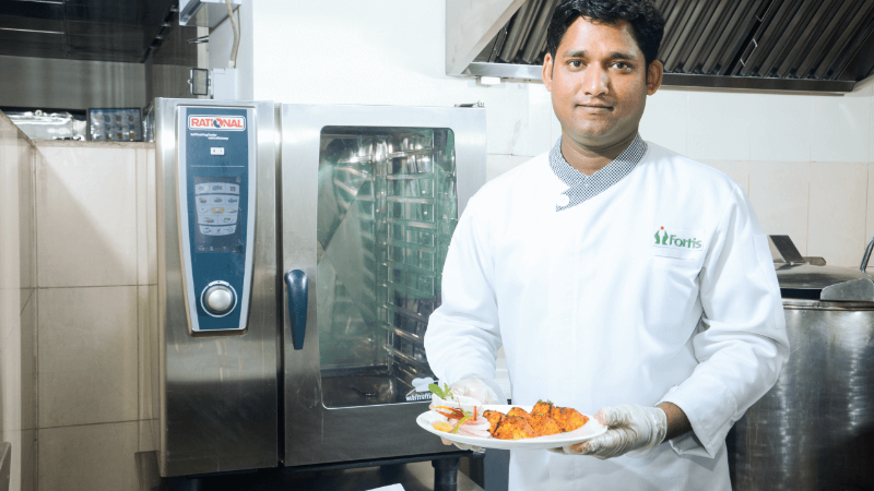 A chef holding a plate of food in front of a Rational oven in a commercial kitchen.