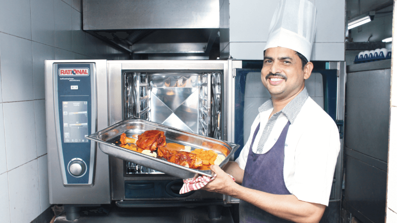 A chef holding a tray of cooked food in front of a Rational oven, smiling and wearing a chef‘s hat.