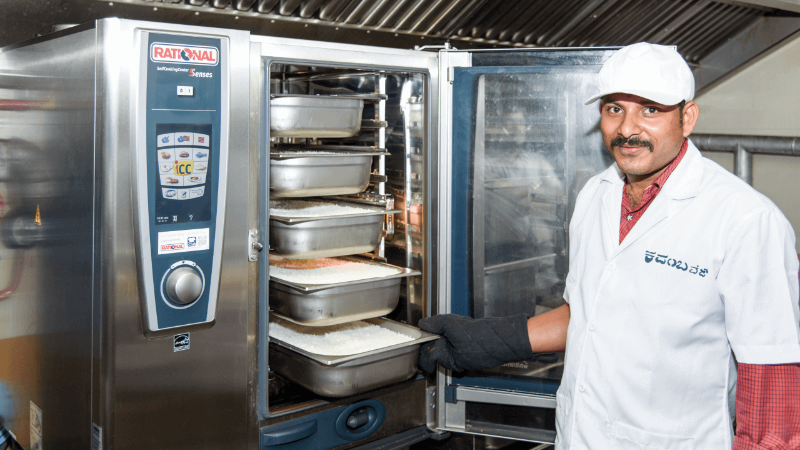 A chef in a white uniform and hat, wearing oven mitts, standing next to a Rational oven with trays of food inside.