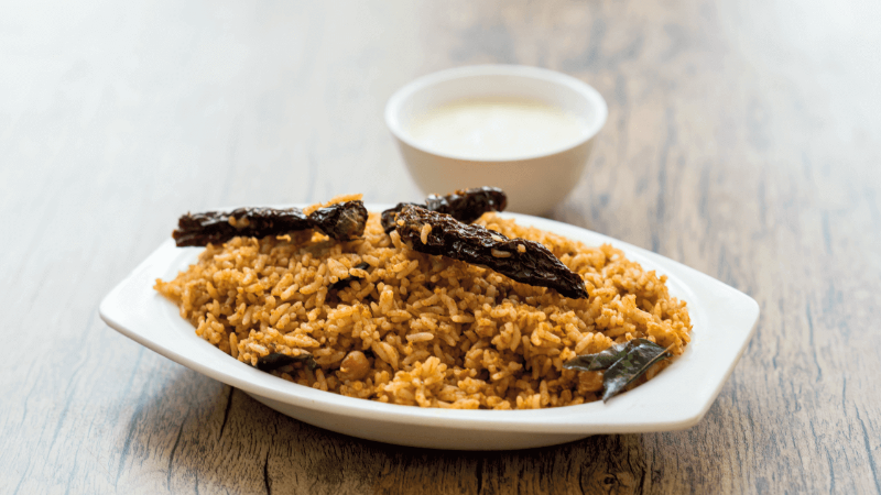A plate of rice with dried chili and leaves, served with a bowl of yogurt on the side.