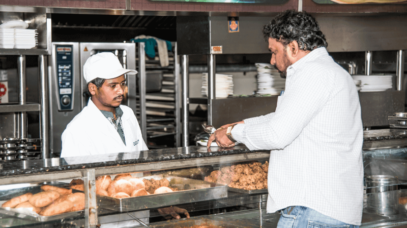 A restaurant employee serving food to a customer at the counter, with trays of food and a Rational oven in the background.