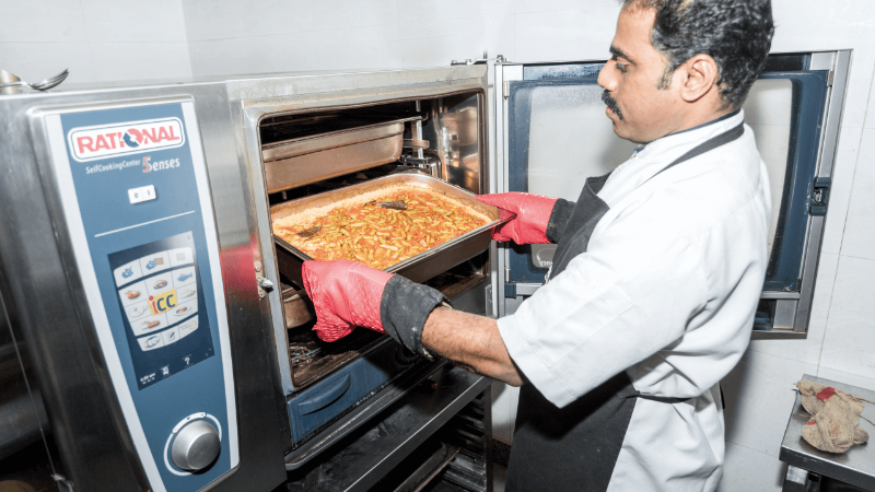A chef wearing red gloves is removing a tray of food from a Rational oven in a kitchen.