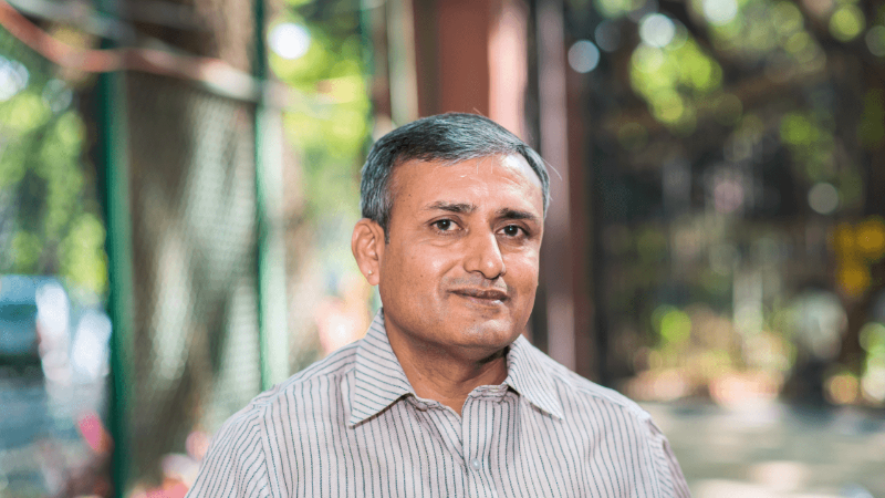 A man with short hair, wearing a striped shirt, stands in front of a green fence, smiling slightly.