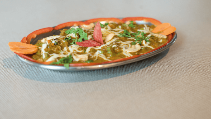 A dish of green curry served with grated cheese, cashews, and garnished with carrot slices and fresh cilantro.