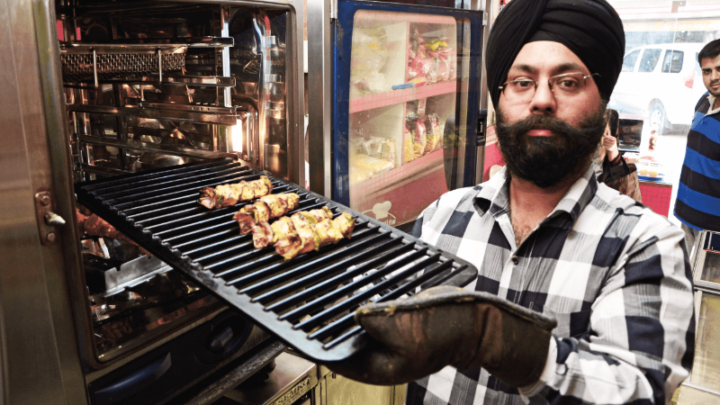 A man wearing a turban holding a tray with grilled skewers in front of a Rational oven, with others in the background.