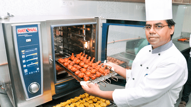 A chef in a white uniform holds a rack of skewered grilled chicken in front of a Rational oven, with a tray of kebabs below.