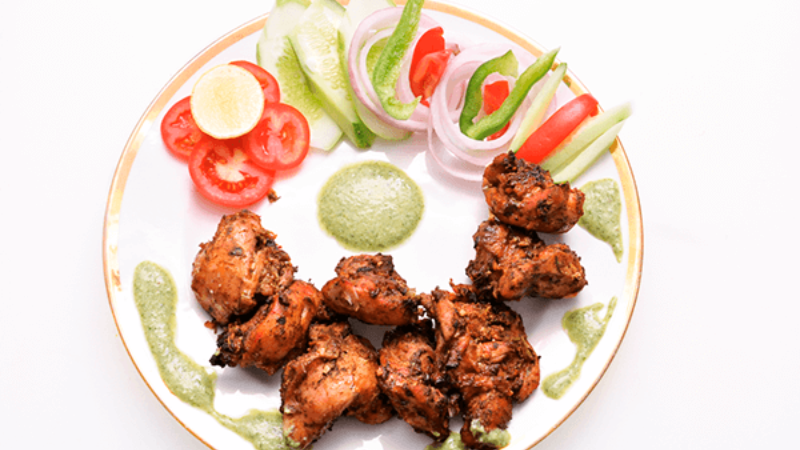 A plate of grilled chicken pieces served with fresh salad, including tomatoes, cucumber, and onions, along with green chutney.