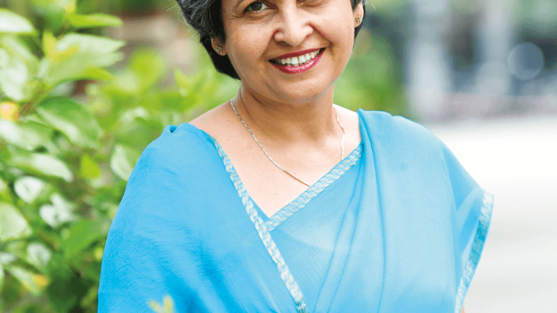 An individual with grey hair wearing a blue sari, smiling outdoors with greenery in the background.
