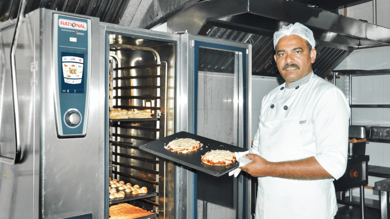 A chef stands next to a Rational oven in a commercial kitchen, holding a tray with two pizzas.