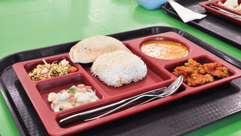 A tray containing a variety of Indian dishes including rice, curry, idli, and sides, served on a green table.