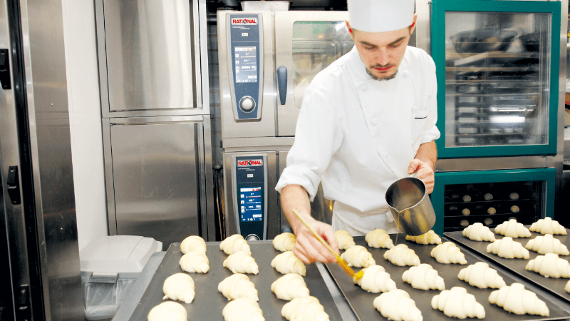  A chef in a white uniform and hat is brushing egg wash onto uncooked croissants arranged on trays in a professional kitchen, with ovens in the background.
