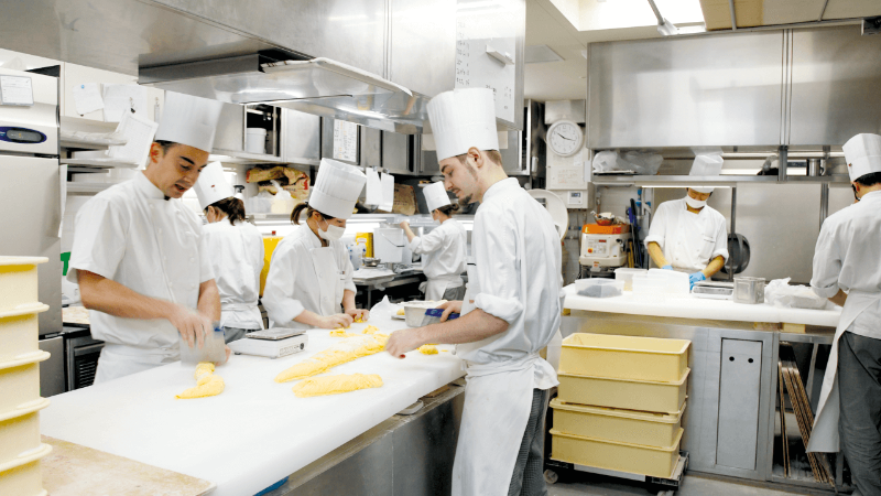 A busy professional kitchen scene with several chefs wearing white uniforms and tall hats, preparing food on a counter with various ingredients and equipment in the background.