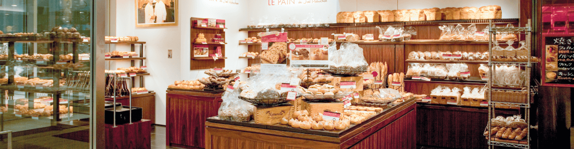 An inviting bakery named "LE PAIN" featuring various types of bread and pastries displayed on wooden shelves, with bright lighting and a photo of the baker on the wall.