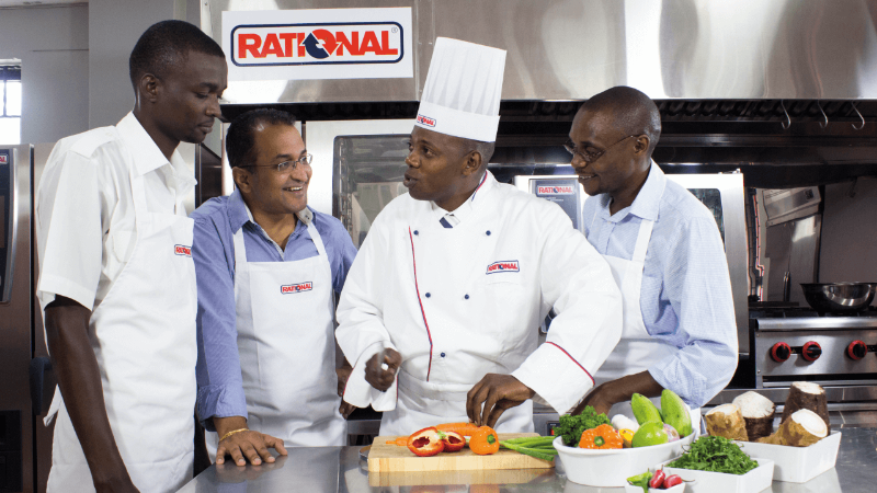 Four people in a kitchen, including a chef in a white uniform and hat, preparing vegetables on a cutting board while others observe.
