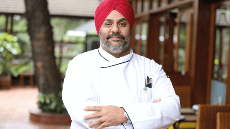 Chef in white uniform and red turban smiling outdoors near a restaurant.