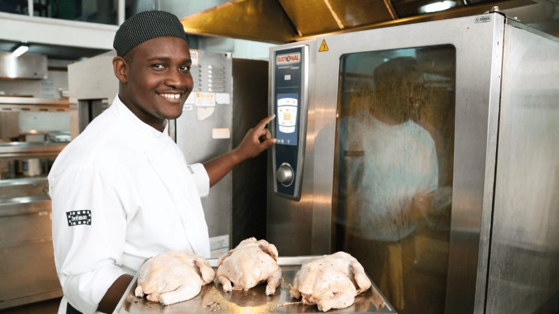Chef smiling while holding raw chickens on a tray next to a Rational combi oven.