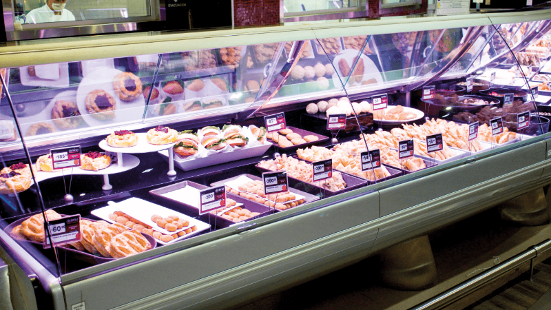 A bakery display case with various pastries and sandwiches, with price tags visible. A staff member is visible behind the counter.