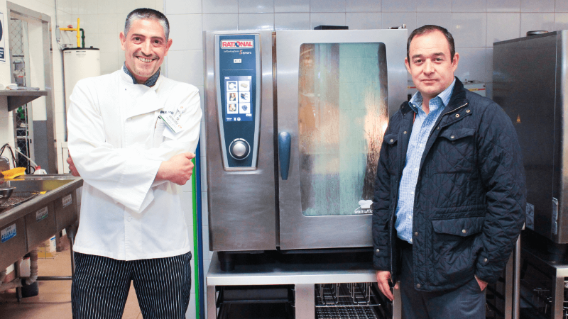 Two men standing next to a Rational oven in a kitchen, smiling and posing for the photo.