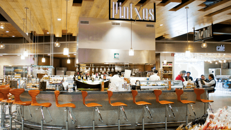 The interior of a modern restaurant with a bar area, chefs working in the kitchen, and customers seated at a table.