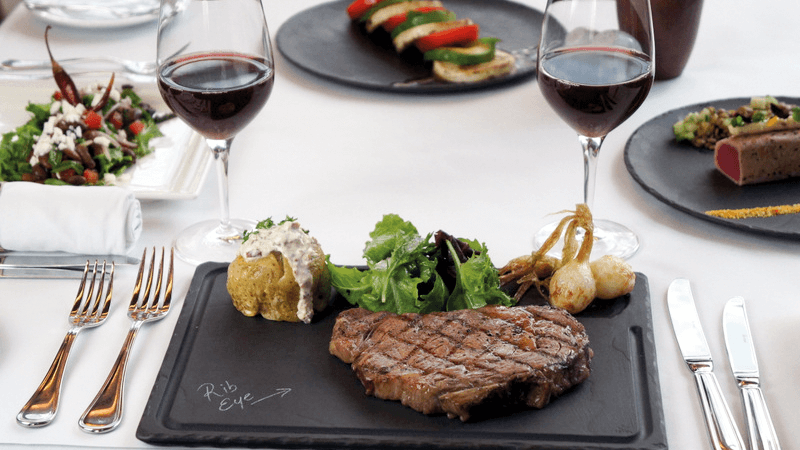 Ein Teller mit gegrilltem Rib-Eye-Steak, Ofenkartoffel, Zwiebeln und Salat, dazu zwei Gläser Rotwein.