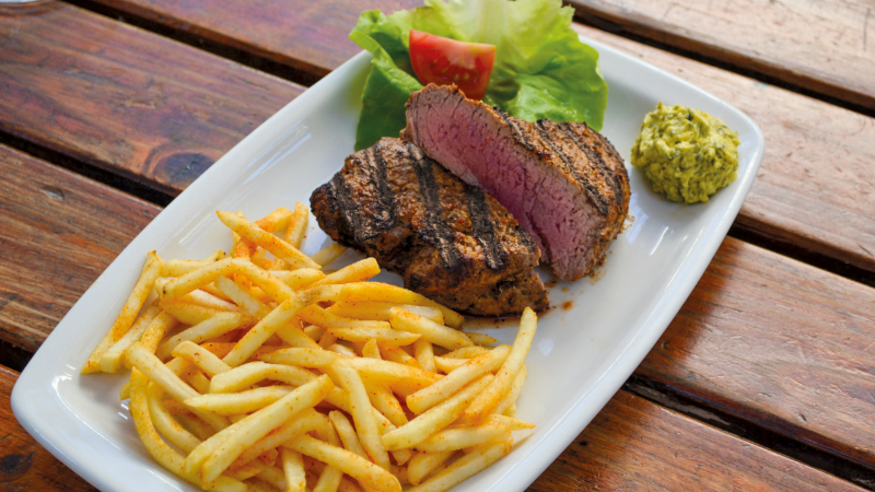 A plate with grilled steak, a side of French fries, a piece of lettuce and tomato, and a dollop of green sauce.