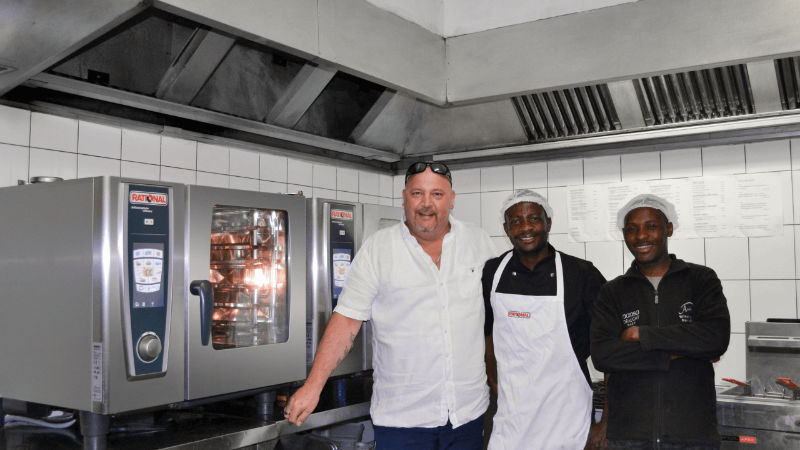 Three men, including a chef in a white shirt and apron, pose in a kitchen with a Rational oven in the background.
