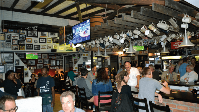 A busy bar with patrons sitting at tables, a television screen showing a game, and mugs hanging from the ceiling, with walls decorated with photos.