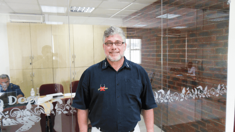 Smiling man wearing a black shirt with the word "Patience" on it, standing in an office setting with glass walls.