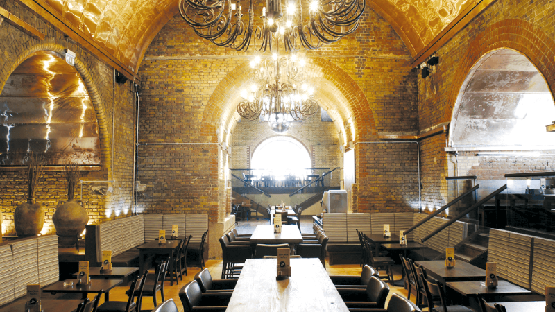 Elegant restaurant interior with brick walls and large chandeliers, featuring wooden tables and chairs.