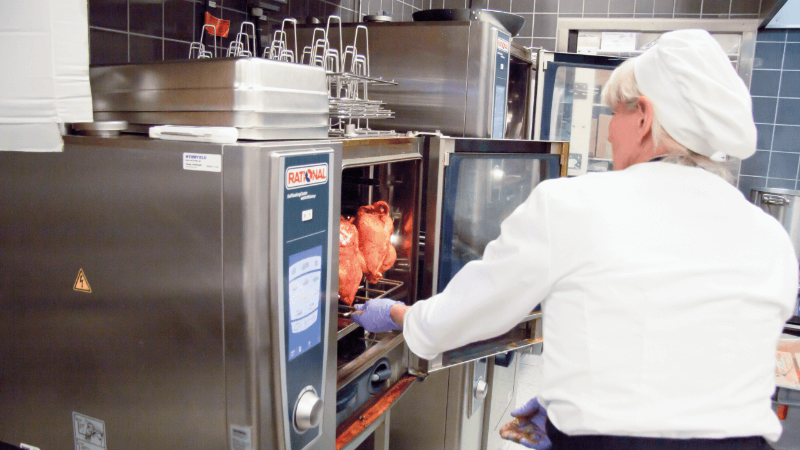 Ein Koch in einer Küche, der einen weißen Hut und Handschuhe trägt und Essen in einen Rational-Ofen legt.