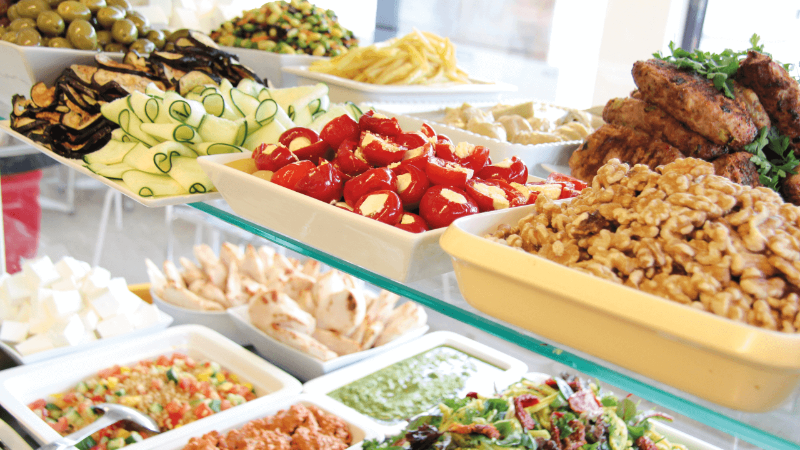 A vibrant display of assorted salads and appetizers, including olives, zucchini, stuffed peppers, and walnuts, arranged beautifully on a glass shelf.