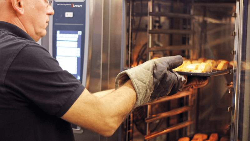 A chef wearing an oven mitt is carefully pulling a tray of cooked food from a Rational oven, showcasing a professional kitchen environment.