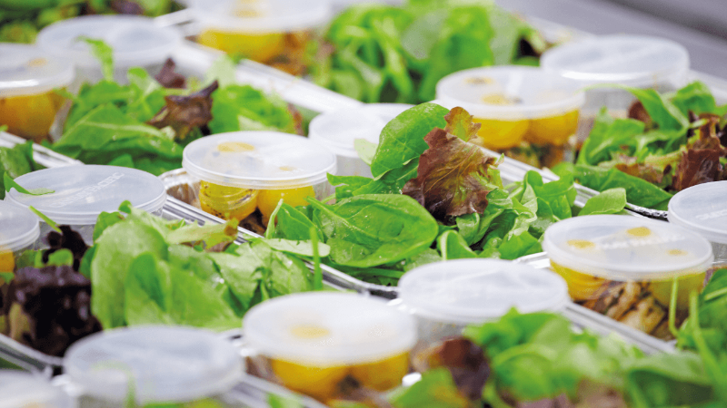 Close-up of multiple containers filled with fresh mixed greens and salad dressing, arranged in a neat row.