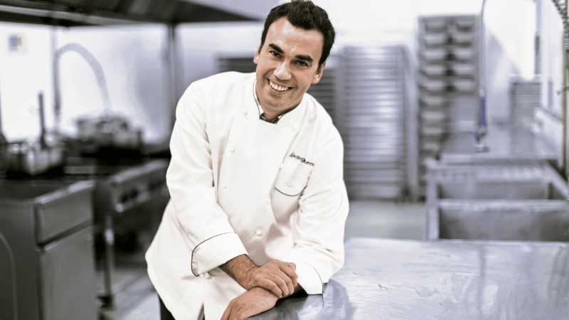 A smiling chef in a white uniform leaning on a shiny metal countertop in a commercial kitchen.