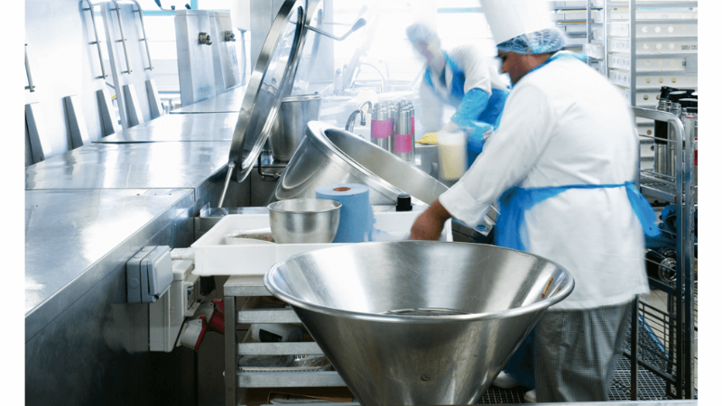 Two chefs in a commercial kitchen working with large stainless steel bowls and pots, creating meals in a busy environment.