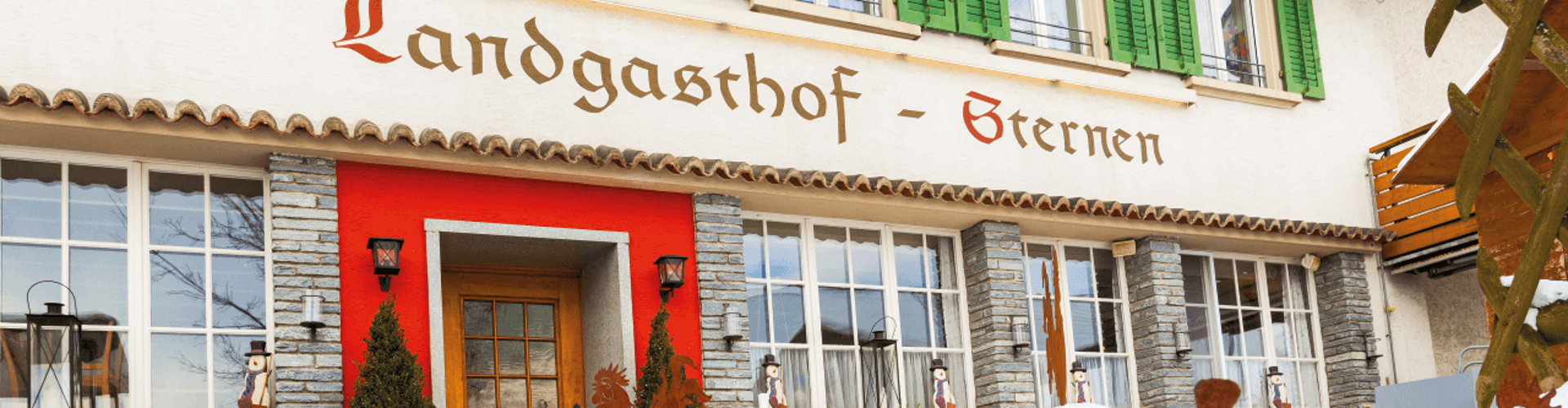 The exterior of a cozy restaurant called "Landgasthof - Sternen" with green shutters, a wooden door, and snow covering the ground.