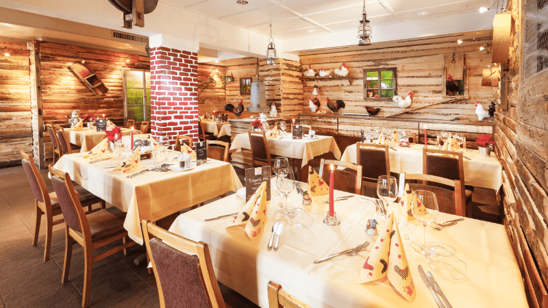 Interior of a cozy restaurant with wooden walls, brick pillars, and tables set with candles and napkins, decorated with rooster figurines.