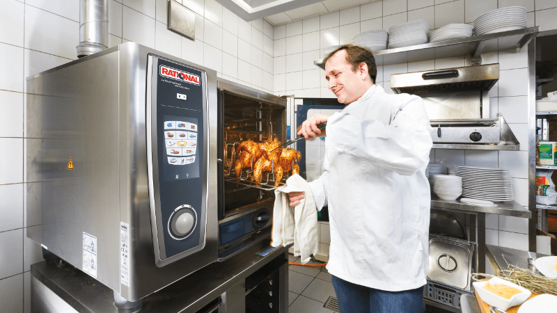 A chef removing roasted chickens from a Rational oven in a kitchen, wearing a white jacket and using a towel.
