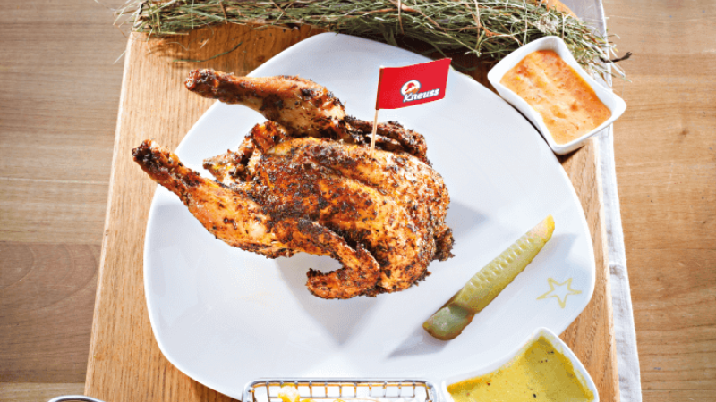 A plate with a roasted chicken, French fries in a small basket, a pickle, a small bowl of sauce, and a sprig of herbs.
