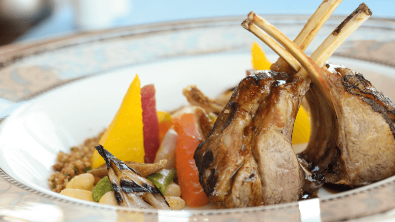Ein Teller mit gegrilltem Lammrippen, Couscous, gegrilltem Gemüse und bunten Zitrusfrüchten.